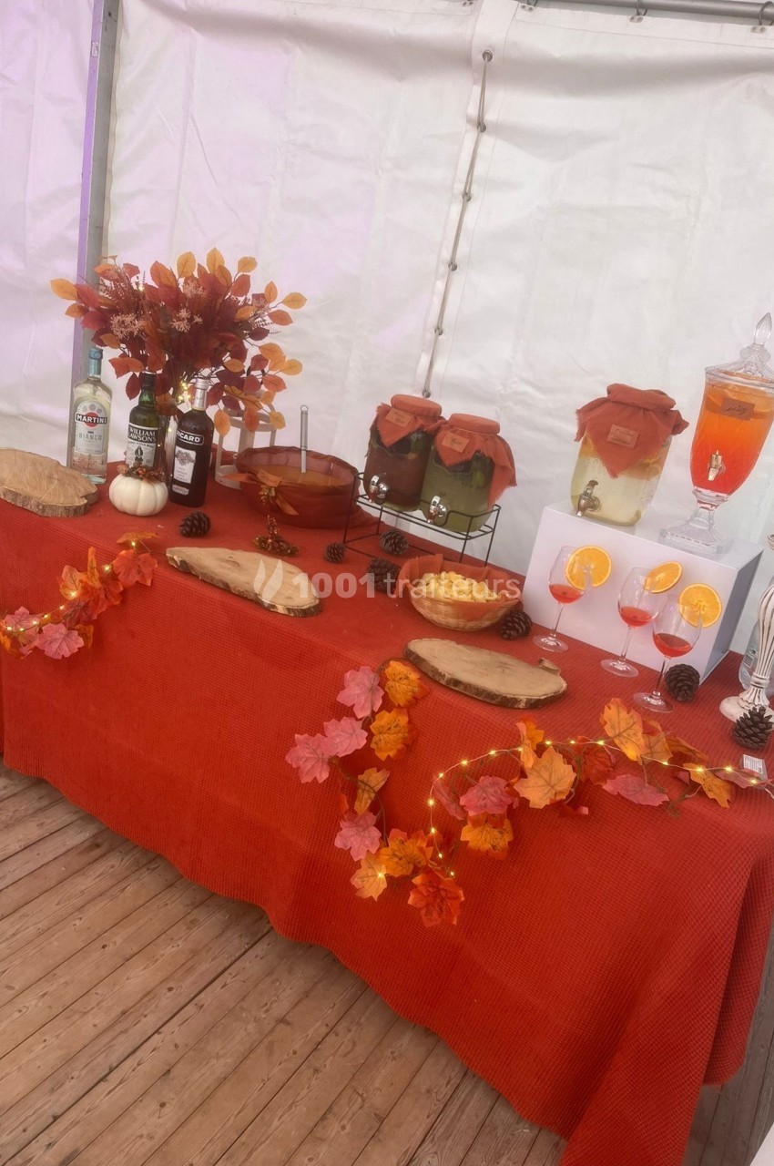 Table décorée aux couleurs de l'automne avec boissons, fruits, fleurs et guirlandes de feuilles sur une nappe rouge.