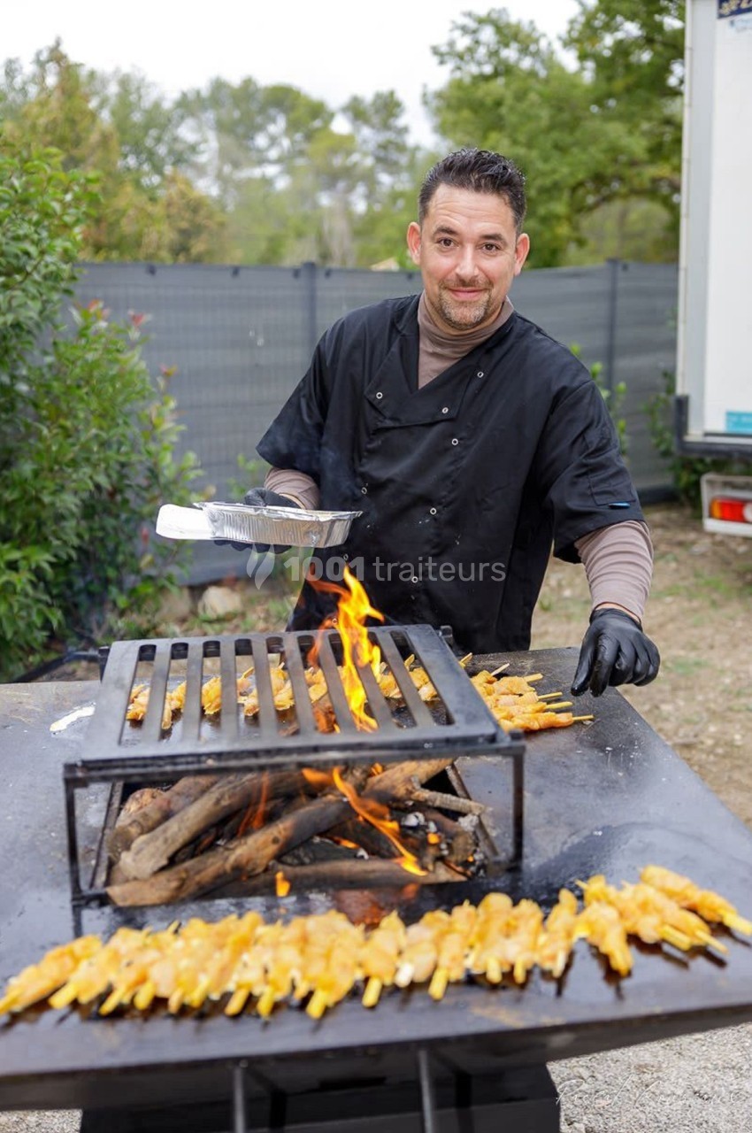 Un cuisinier prépare des brochettes de viande sur un barbecue en plein air avec des flammes vives.