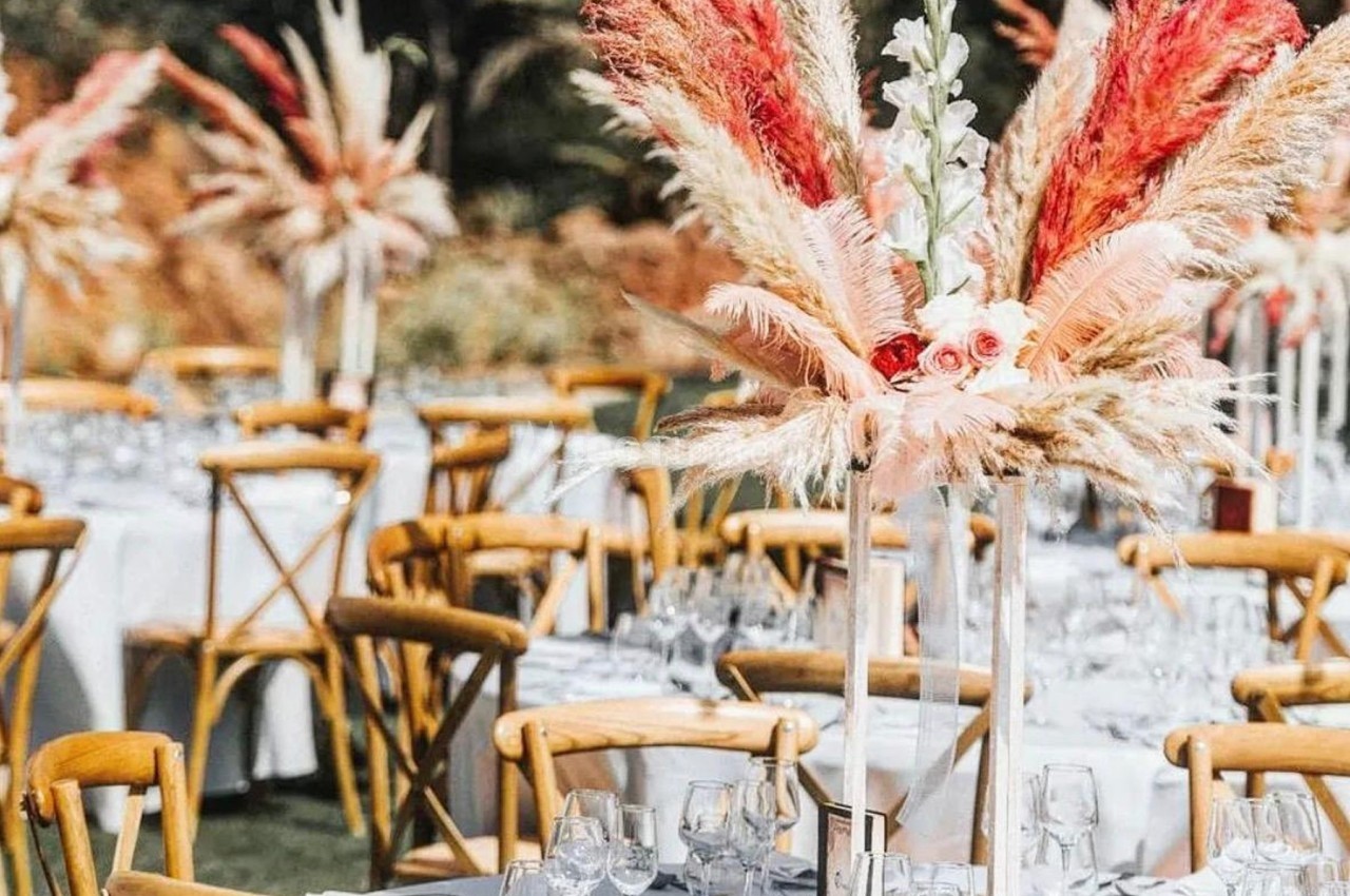 Tables de réception en extérieur décorées de nappes blanches, verres, et centres de table avec fleurs et pampas colorées.