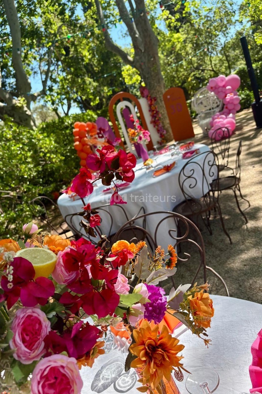 Tables décorées avec des nappes blanches, fleurs colorées et ballons, dans un jardin ensoleillé entouré de verdure.