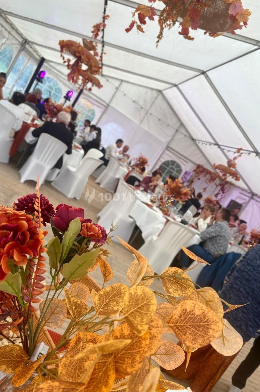 Des invités assis à des tables décorées de fleurs et feuillages d'automne dans une grande tente lumineuse.
