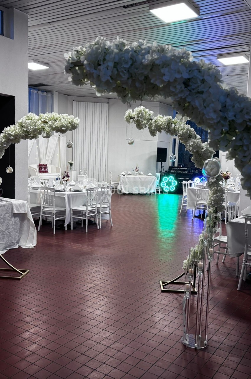 Salle de réception décorée avec des arches fleuries blanches, des tables dressées et un éclairage coloré en arrière-plan.