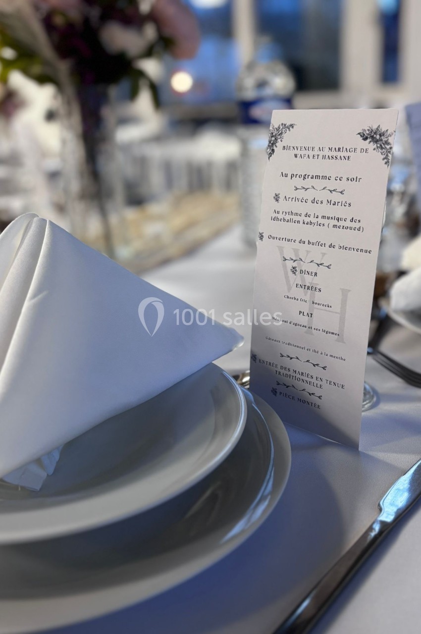 Serviette pliée en pointe posée sur une assiette, avec un menu de mariage imprimé en arrière-plan sur une table dressée.