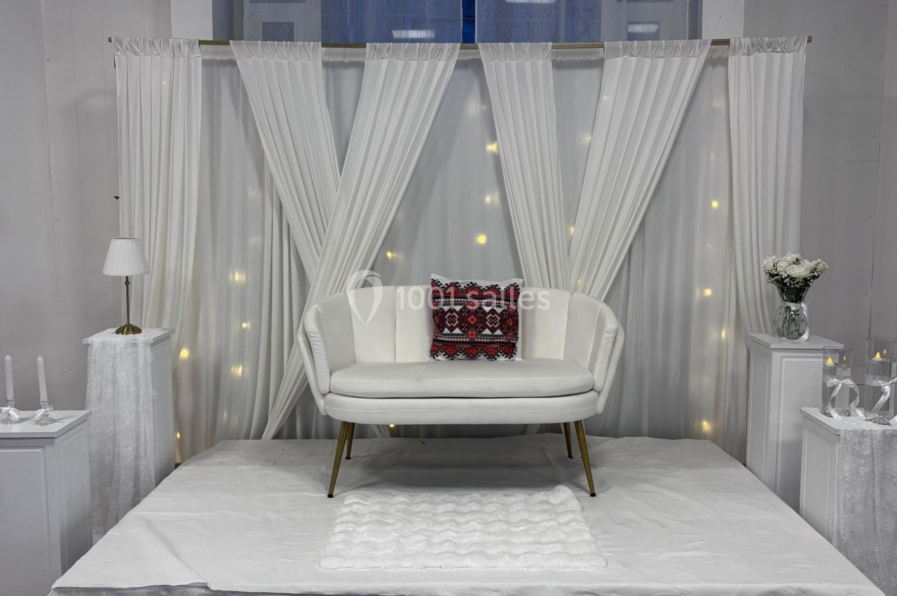 Canapé blanc avec coussin rouge à motifs, placé sur une estrade décorée de rideaux blancs et lumières tamisées.