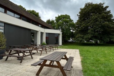 Tables de pique-nique en bois sur une terrasse en béton, entourées de pelouse et d'arbres sous un ciel nuageux.