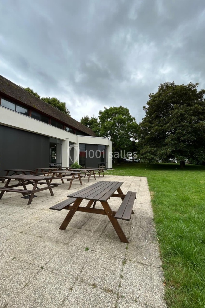 Tables de pique-nique en bois sur une terrasse pavée, entourées de pelouse et d'arbres sous un ciel nuageux.