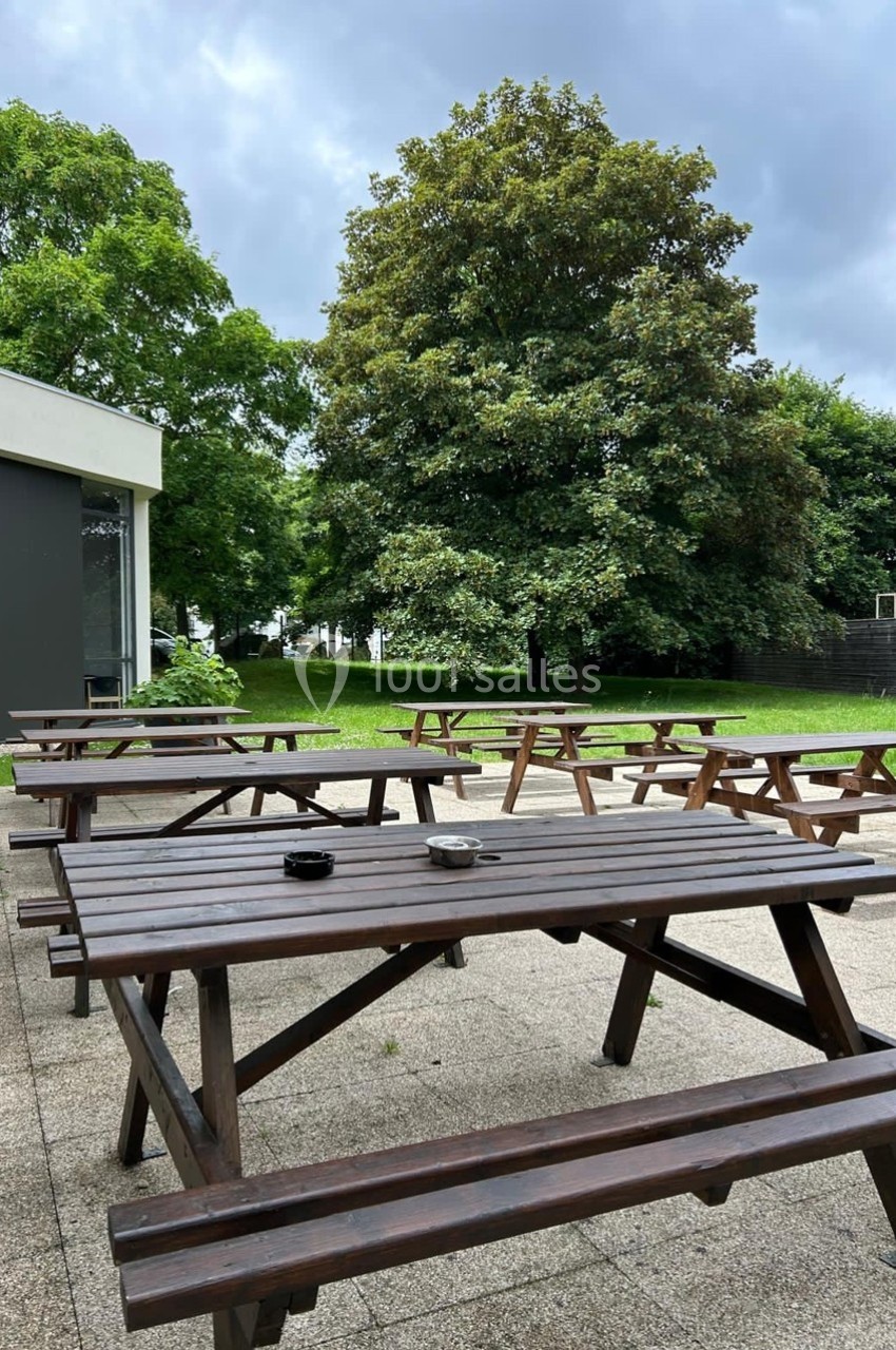 Tables de pique-nique en bois sur une terrasse en béton, entourées de pelouse et d'arbres sous un ciel nuageux.