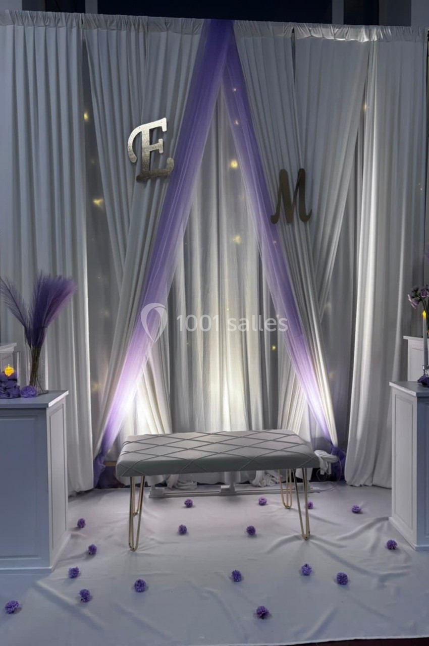 Décor avec rideaux blancs et violets, lettres ’E’ et ’M’, banc blanc, lumières tamisées et pétales au sol.