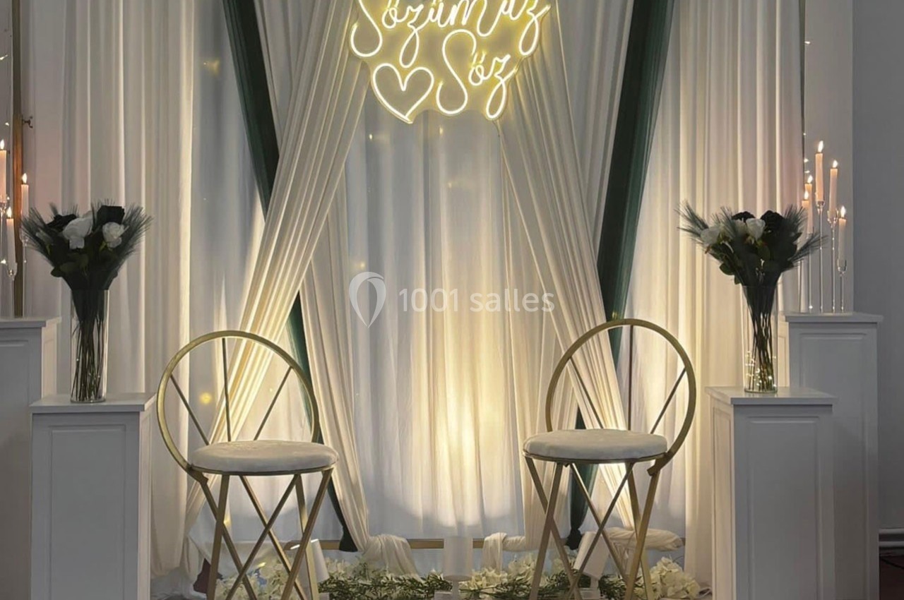 Deux chaises dorées devant un rideau blanc décoré de fleurs, bougies et une inscription lumineuse.