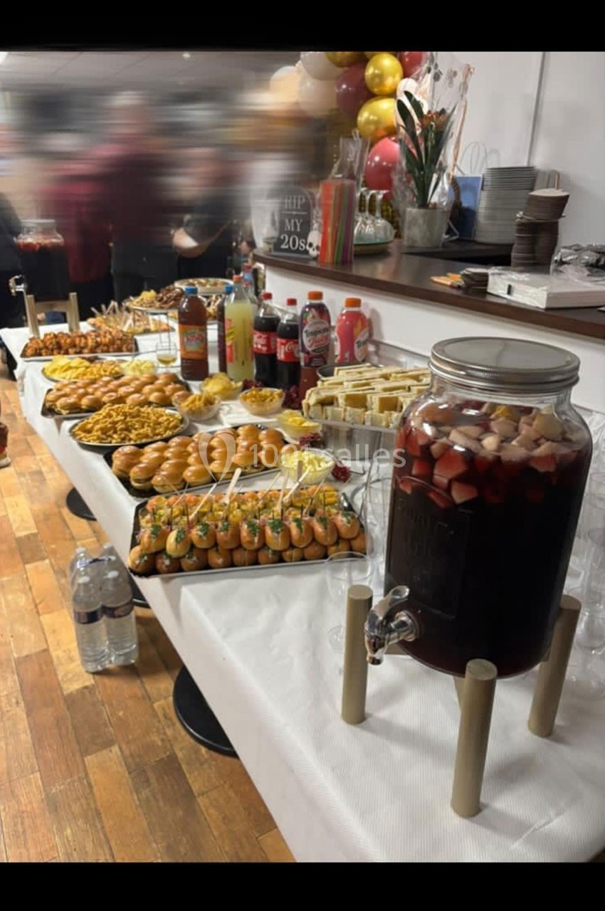 Buffet varié avec boissons, mini-sandwichs, amuse-bouches et sangria dans une grande jarre sur une table en bois.