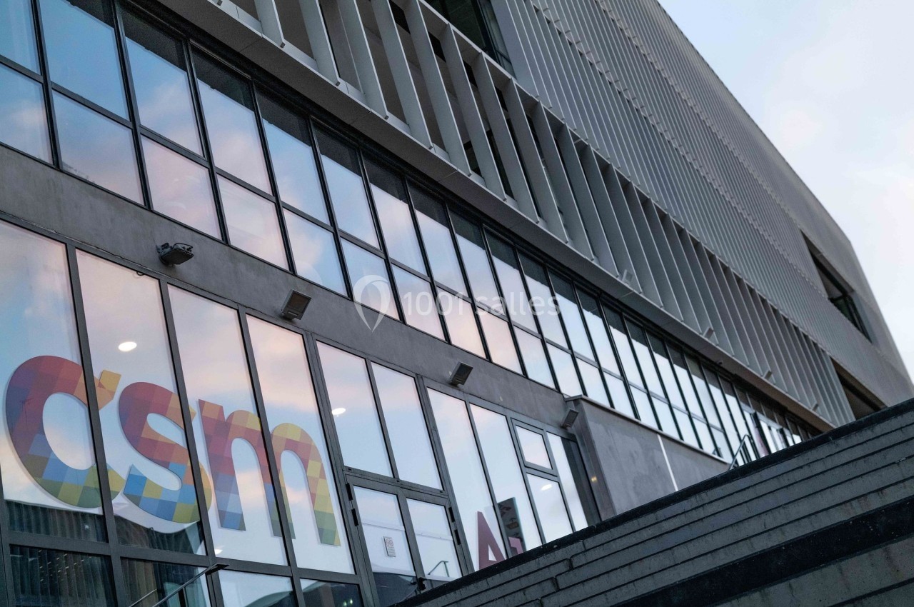 Façade moderne d'un bâtiment avec de grandes baies vitrées reflétant le ciel, et un logo coloré visible sur une vitre.