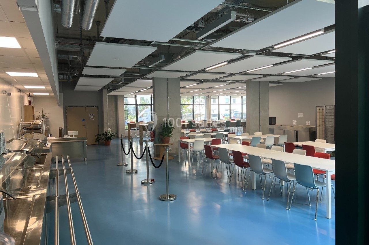 Salle de restauration lumineuse avec tables et chaises colorées, sol bleu et grandes fenêtres donnant sur l'extérieur.