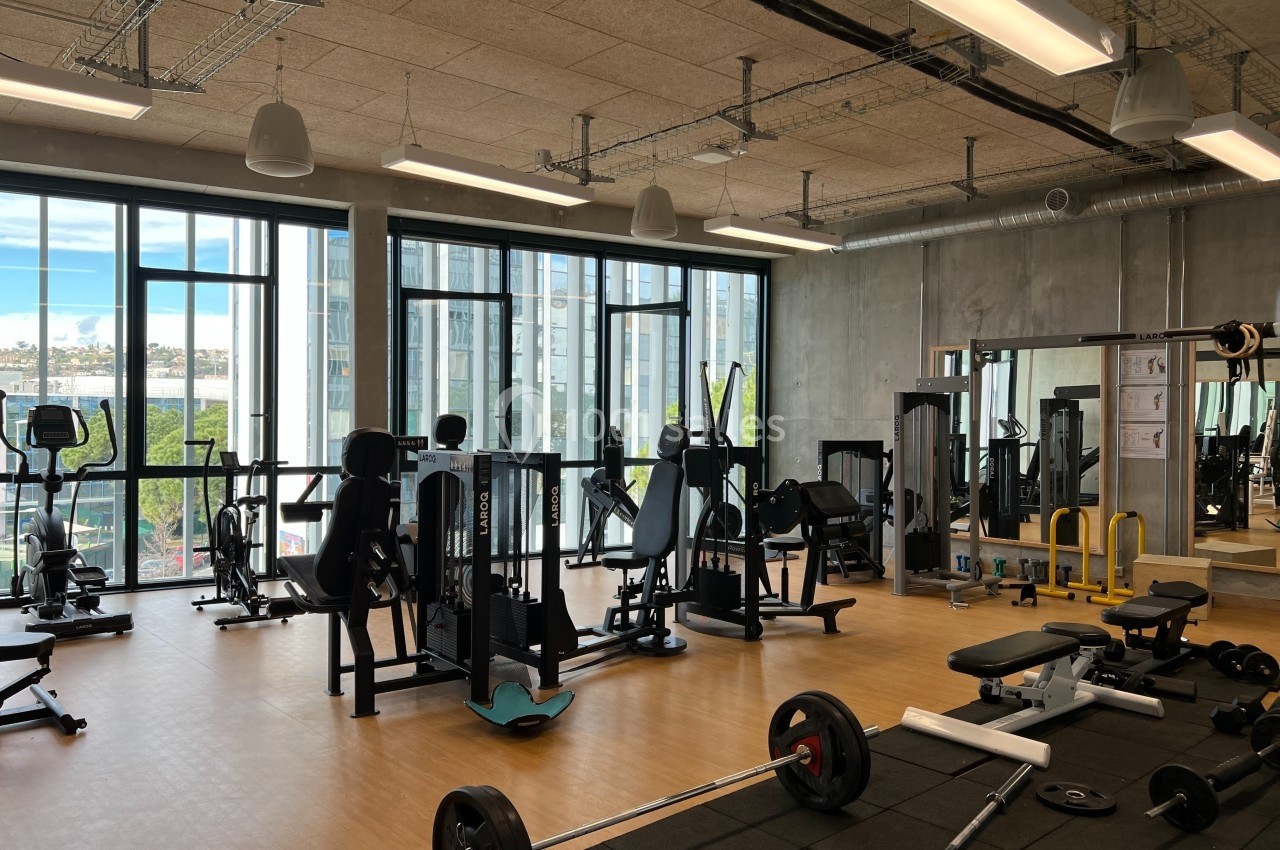Salle de sport lumineuse avec équipements de musculation et de cardio, grandes fenêtres donnant sur l'extérieur.