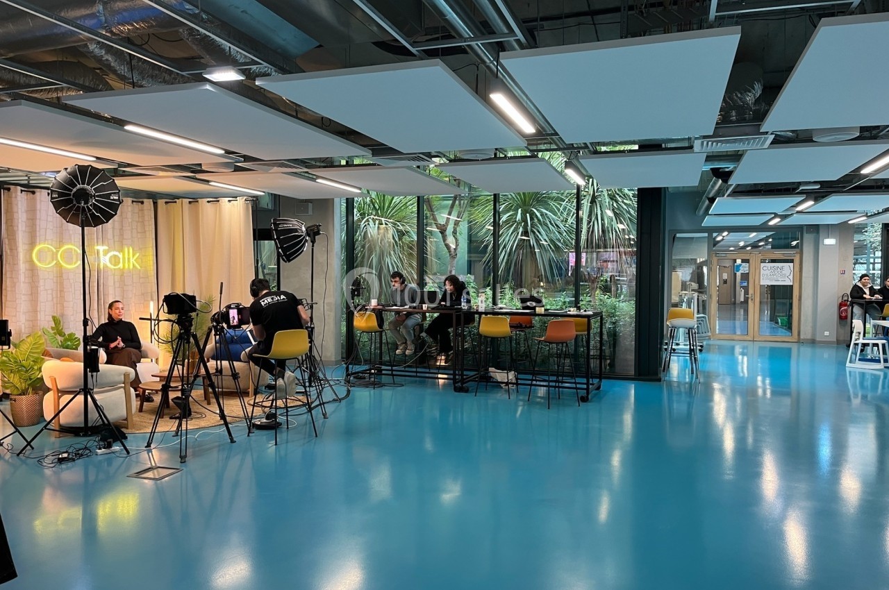 Espace moderne avec tournage en cours, éclairages et caméras, et personnes travaillant sur des ordinateurs en arrière-plan.