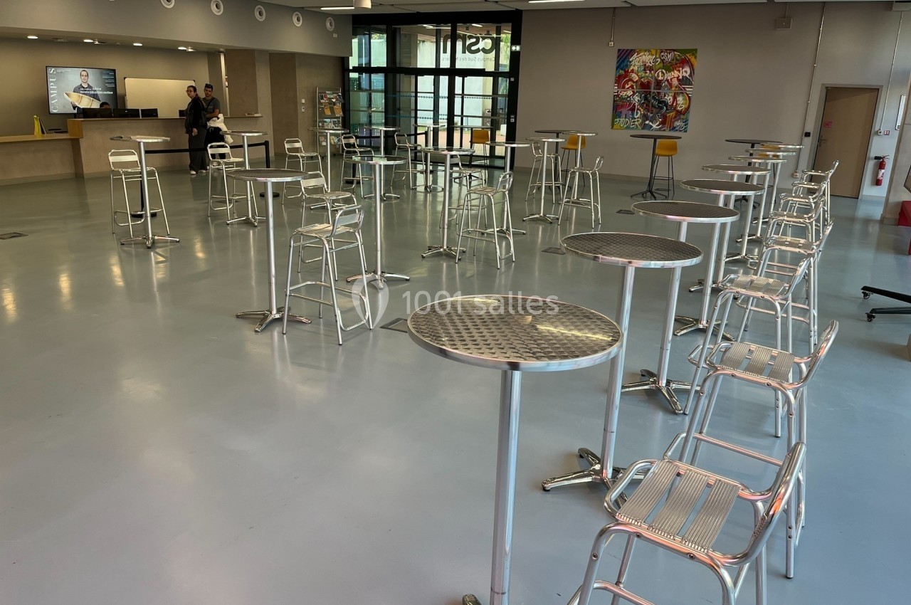 Salle lumineuse avec tables hautes et tabourets métalliques, comptoir d'accueil et tableau coloré en arrière-plan.