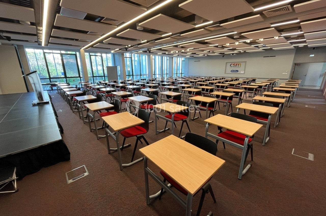 Salle de conférence lumineuse avec des rangées de tables et chaises rouges, équipée d'une scène et de grandes fenêtres.