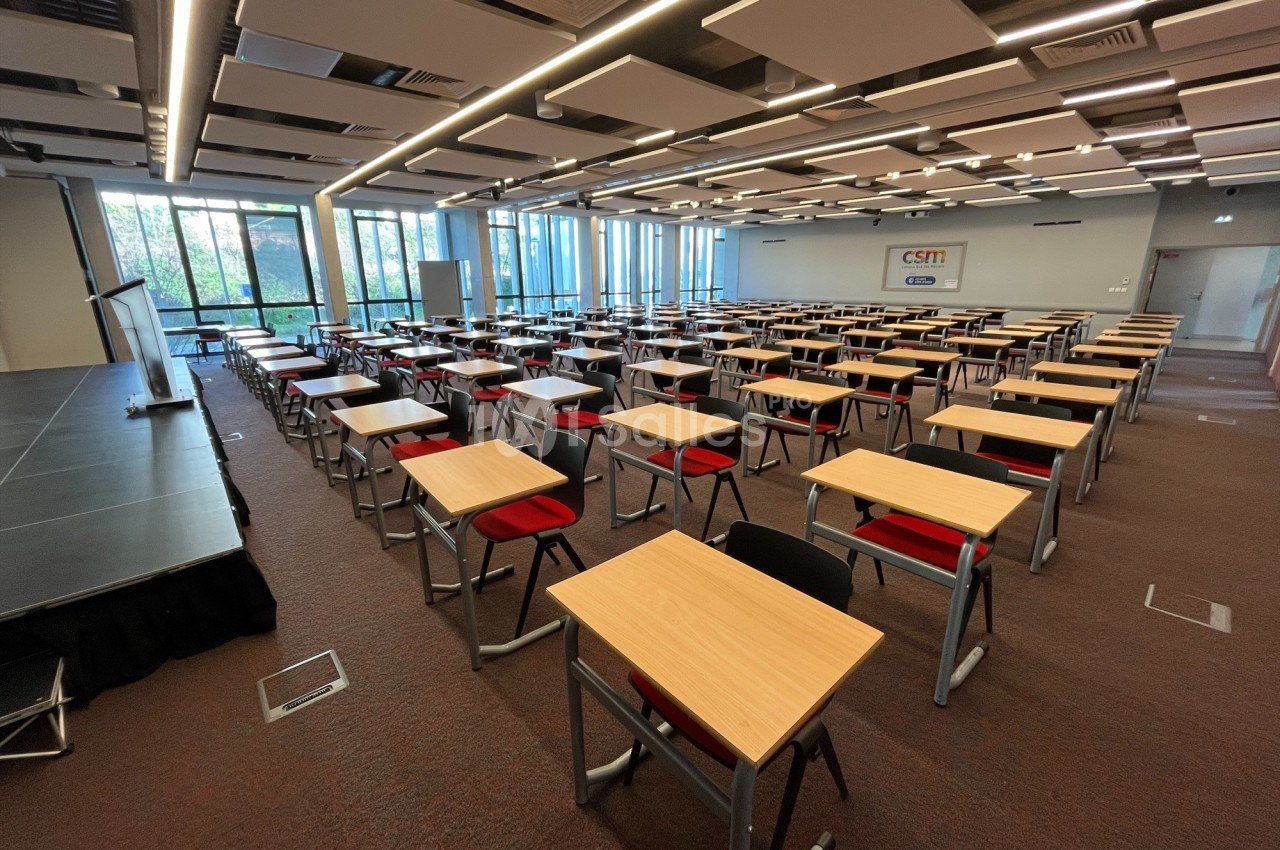Salle de conférence lumineuse avec des rangées de tables et chaises rouges, équipée d'une scène et de grandes fenêtres.