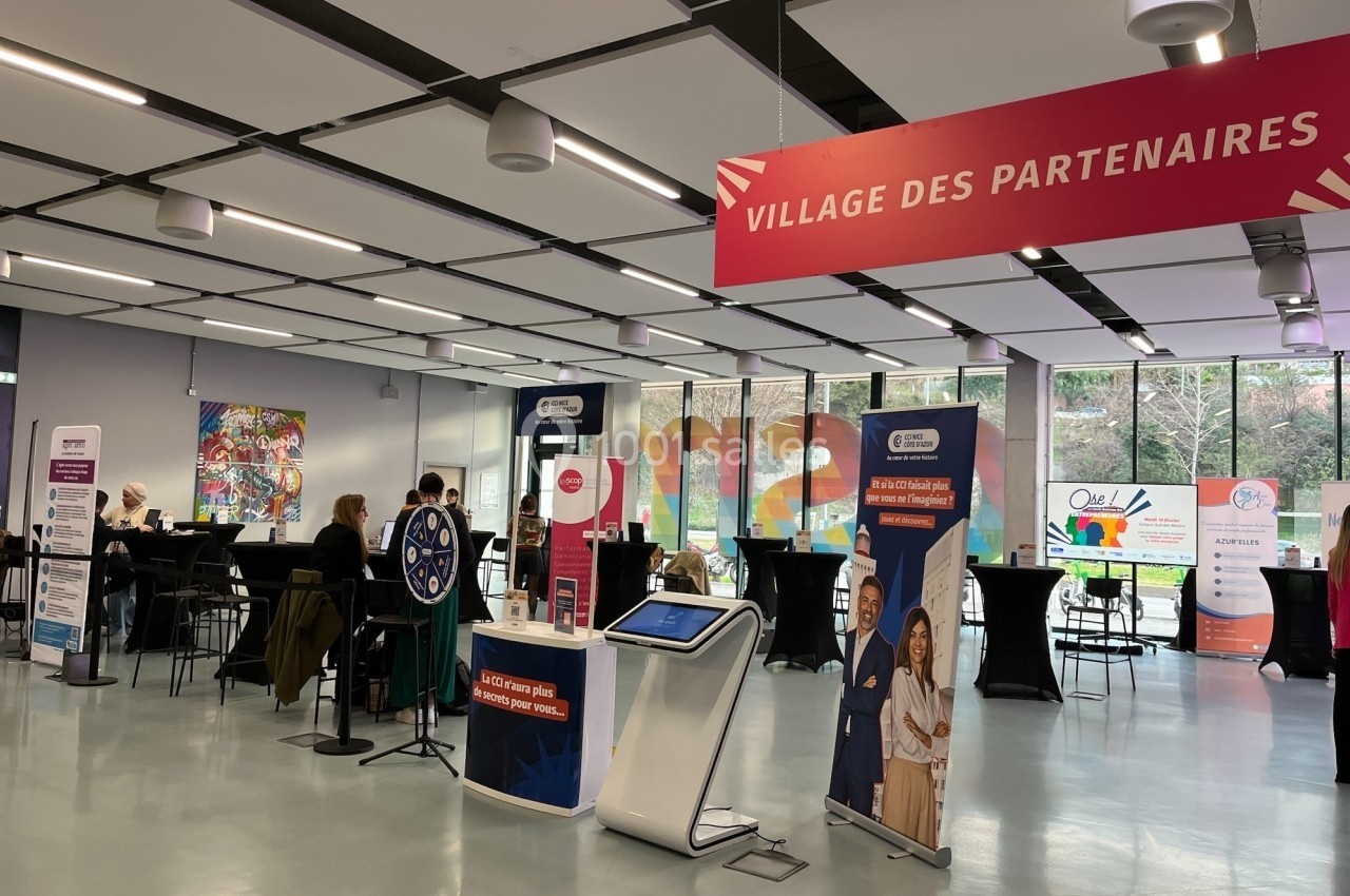 Espace intérieur avec des stands d'information sous une bannière ’Village des partenaires’, quelques personnes présentes.