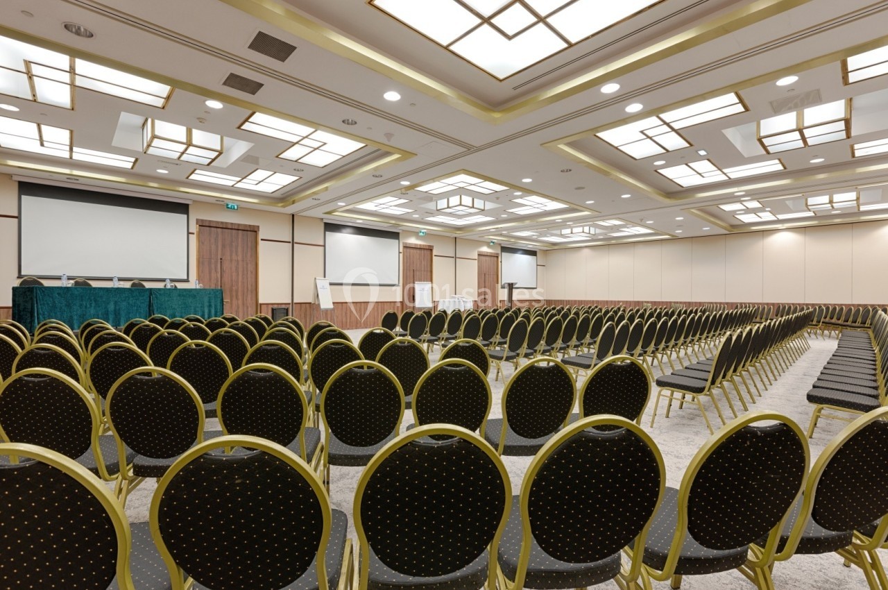 Salle de conférence vide avec des rangées de chaises noires et dorées, trois écrans et un éclairage au plafond.