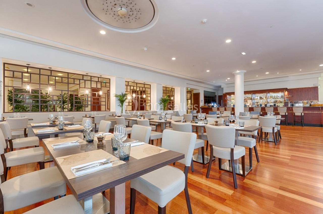 Salle de restaurant lumineuse avec tables en bois, chaises rembourrées et un bar en arrière-plan.