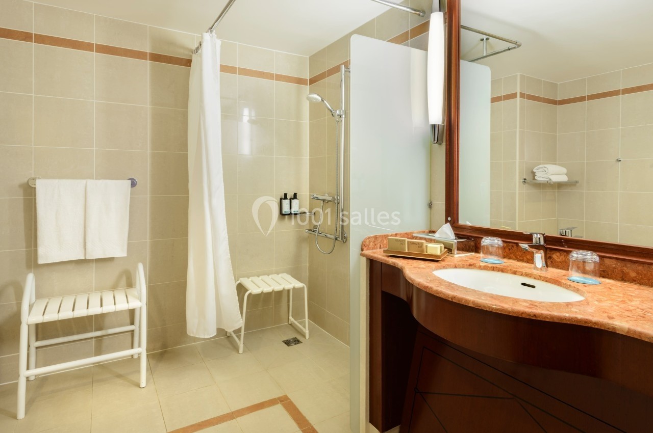 Salle de bain accessible avec douche à l'italienne, rideau, lavabo en marbre et tabourets blancs.