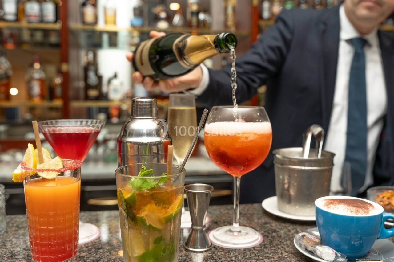 Un barman en costume verse du champagne dans un verre, entouré de cocktails colorés sur un comptoir de bar.