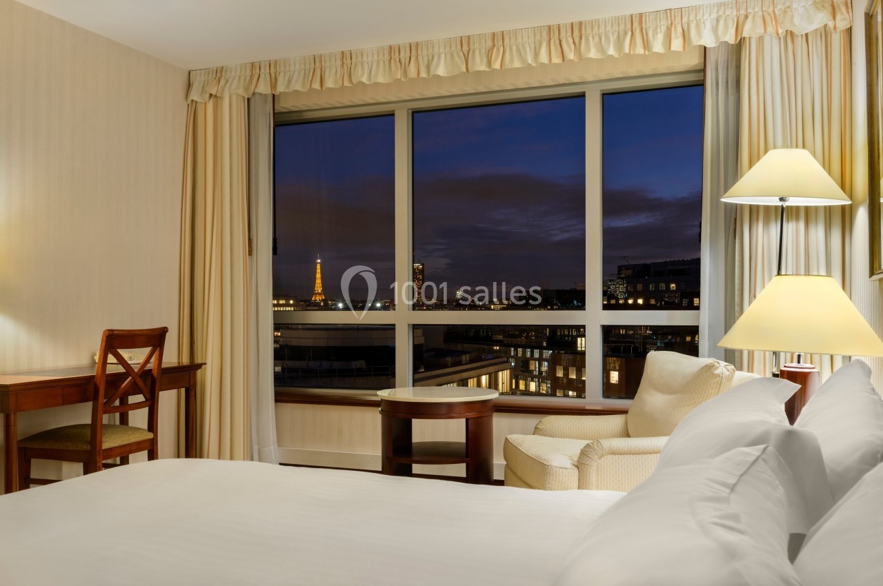 Chambre d'hôtel avec un lit, un bureau, un fauteuil et une vue nocturne sur la Tour Eiffel à travers une grande fenêtre.