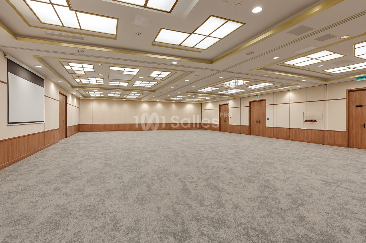 Grande salle vide au sol en moquette grise, plafond à caissons lumineux bordés de dorure, murs crème et boiseries, écran de projection sur la gauche.