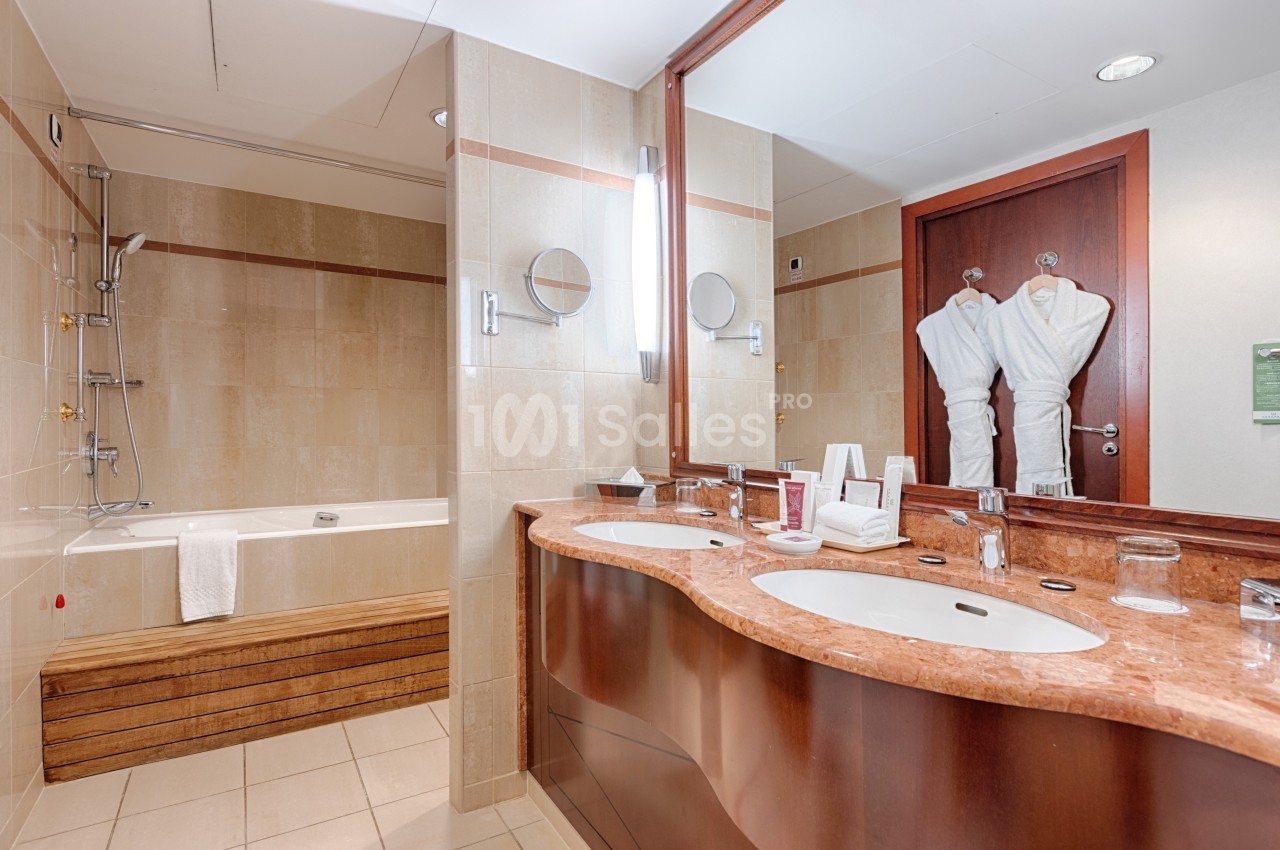 Salle de bain en marbre beige avec double vasque en bois sombre, baignoire, douche et deux peignoirs blancs suspendus près de la porte.