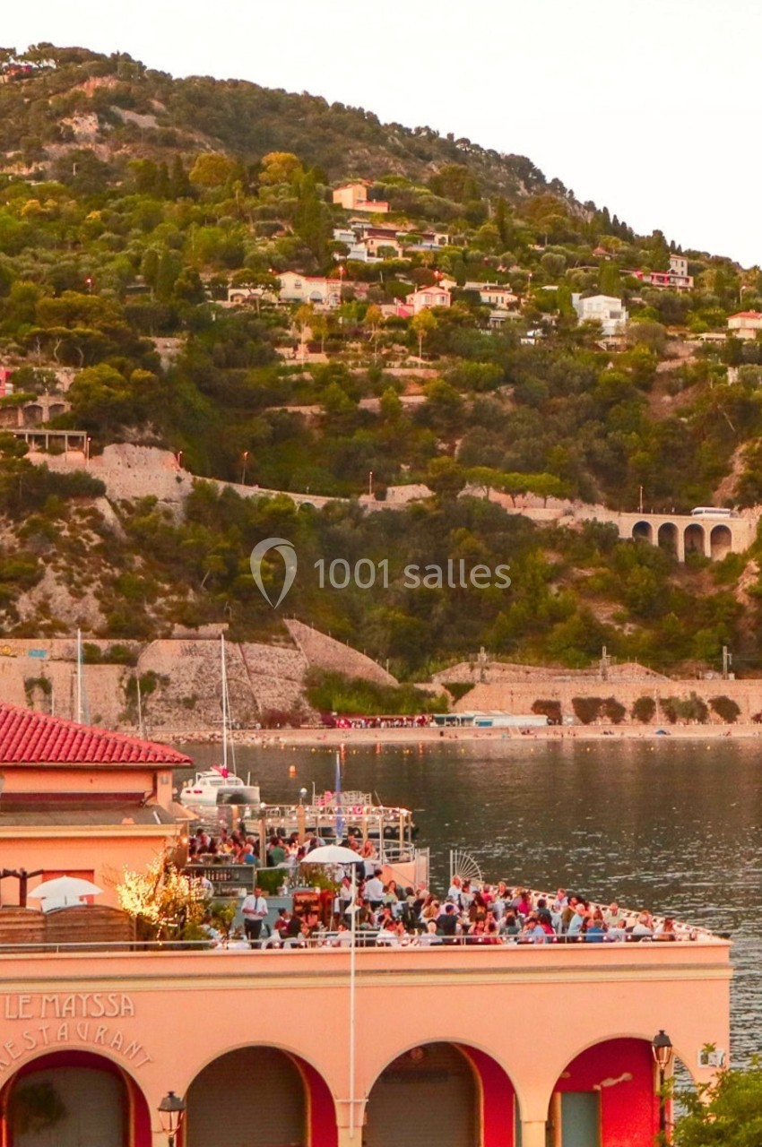 Vue d'une terrasse animée en bord de mer, entourée de collines verdoyantes et de maisons surplombant l'eau.