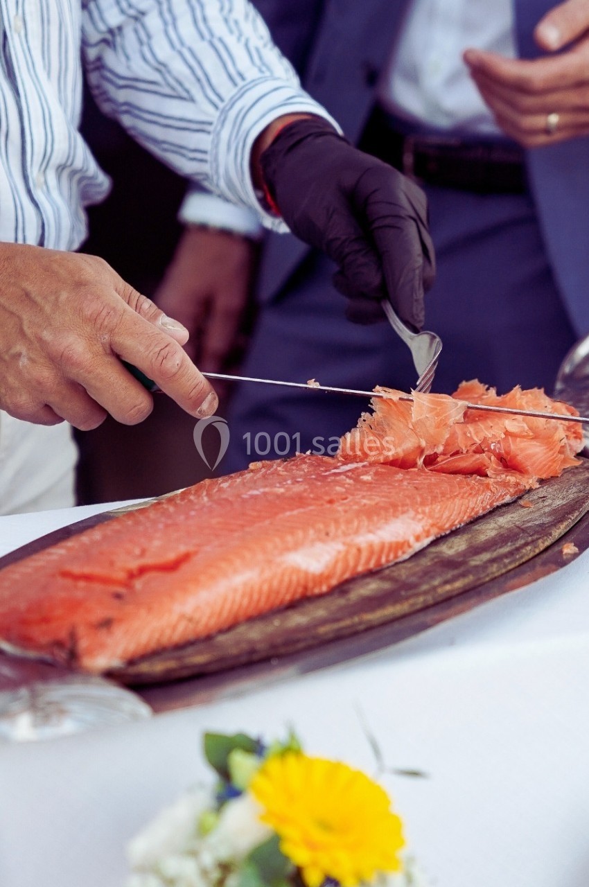 Un homme tranche un filet de saumon frais sur une planche en bois, avec une table décorée en arrière-plan.