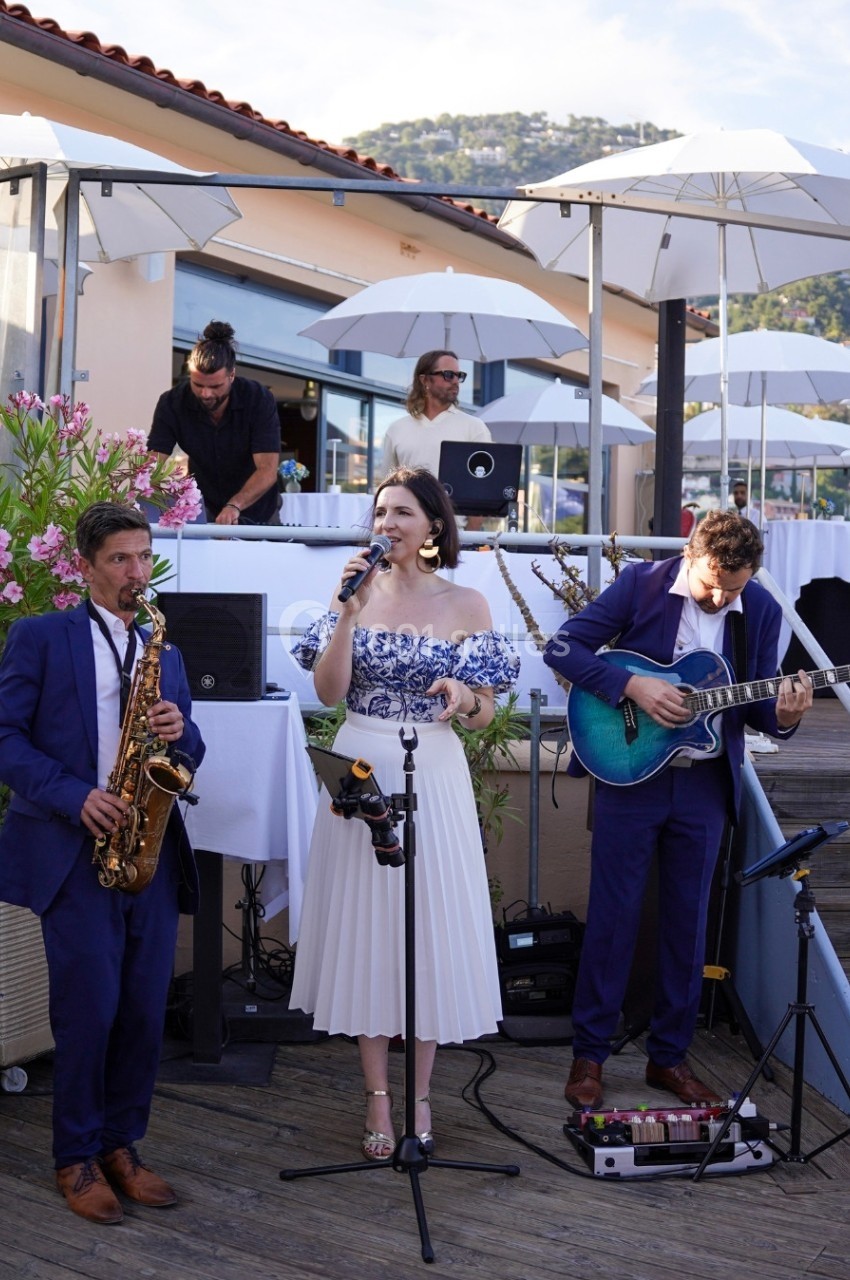 Un groupe de musiciens joue en extérieur sur une terrasse, avec une chanteuse, un saxophoniste et un guitariste.