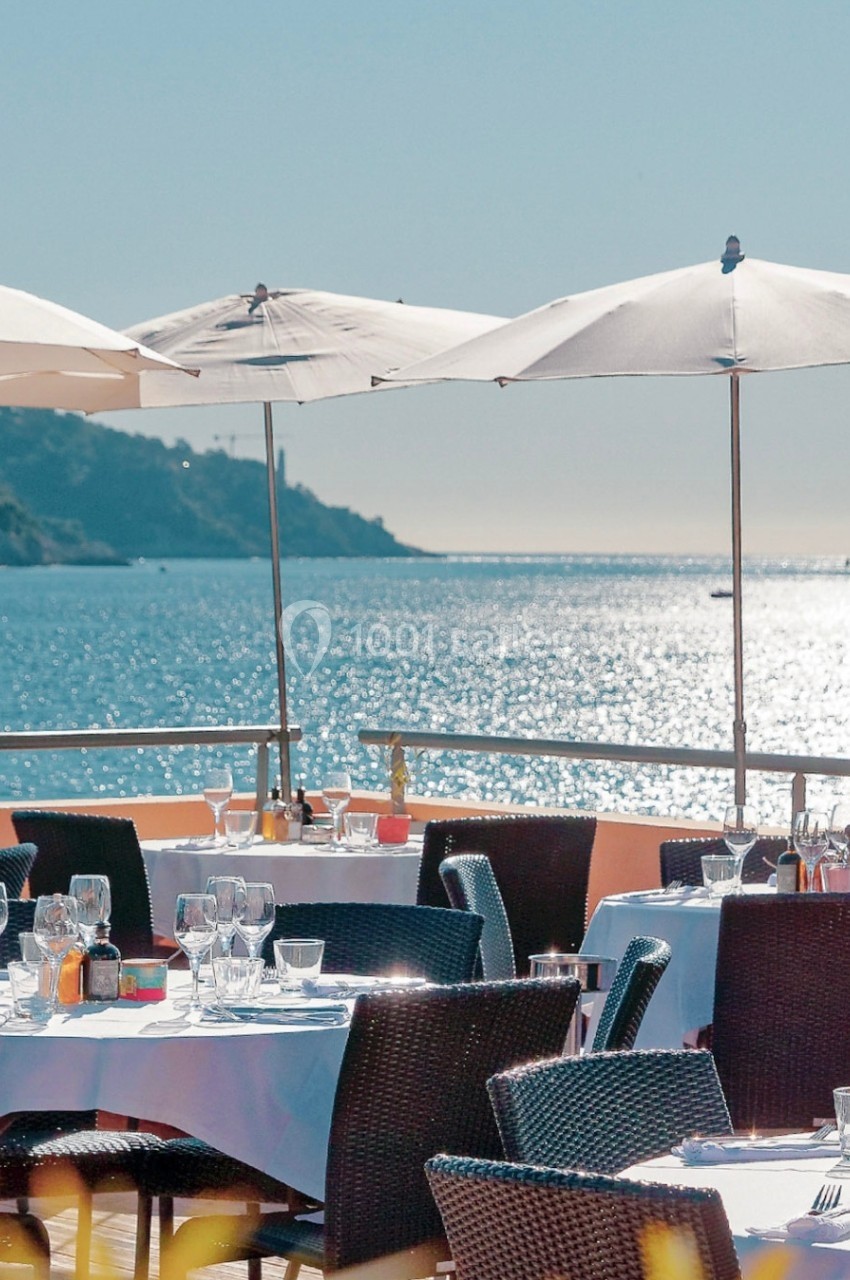 Terrasse en bord de mer avec tables dressées, chaises en osier et parasols sous un ciel ensoleillé.