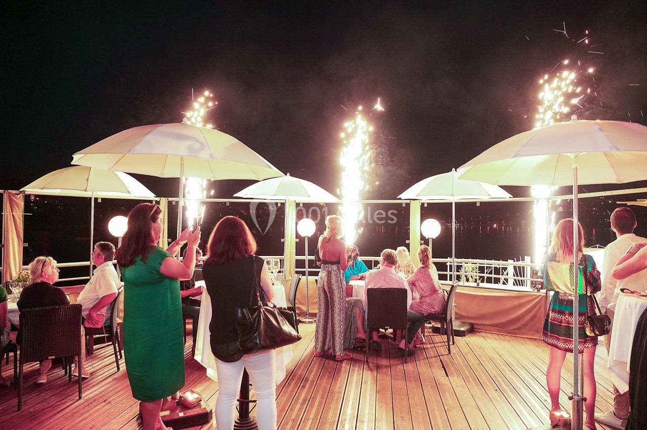 Des personnes dînent en terrasse de nuit, entourées de parasols et de gerbes d'étincelles illuminant la scène.