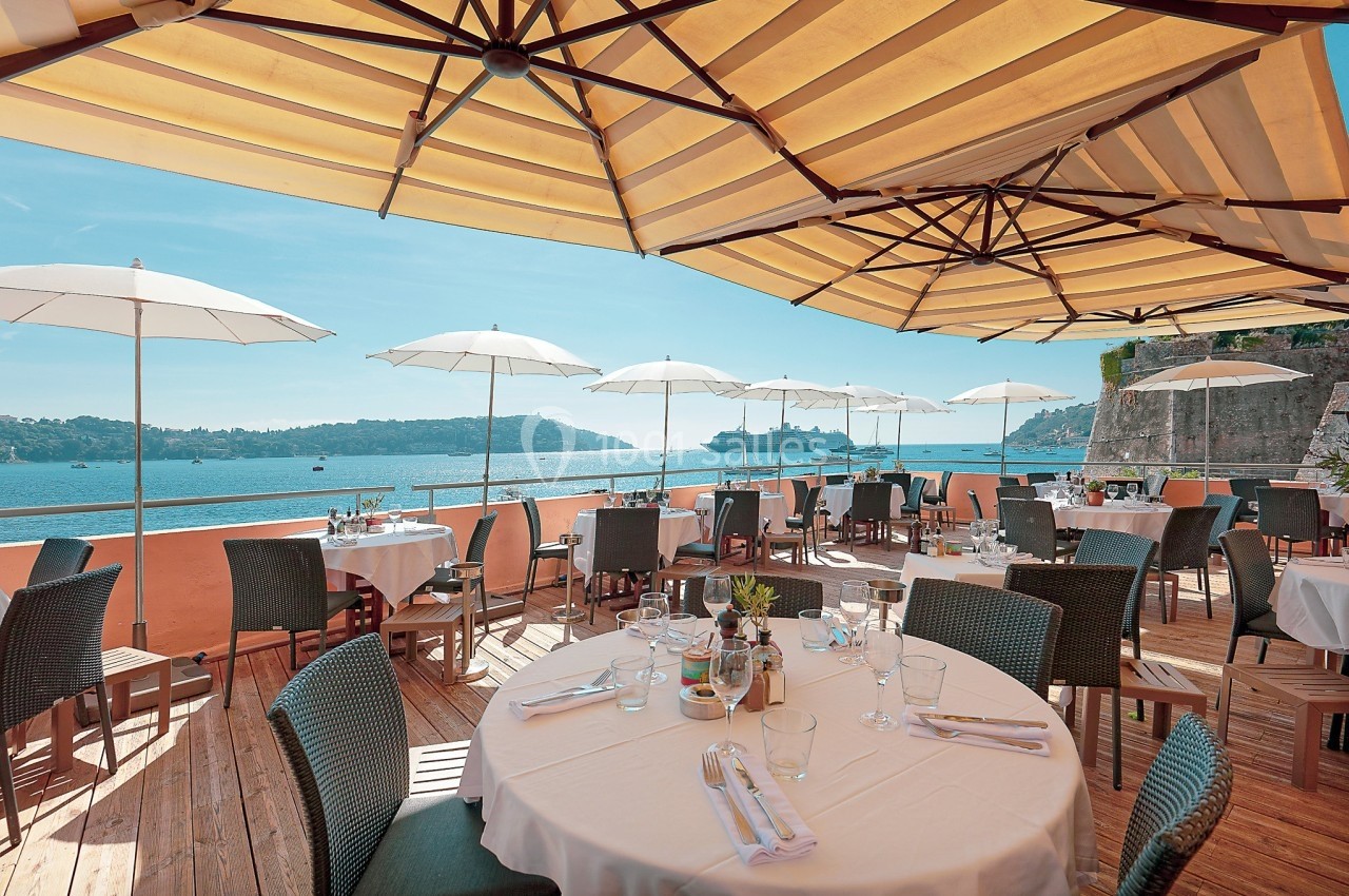Terrasse d'un restaurant avec tables dressées, vue sur la mer, parasols blancs et grandes toiles d'ombrage.