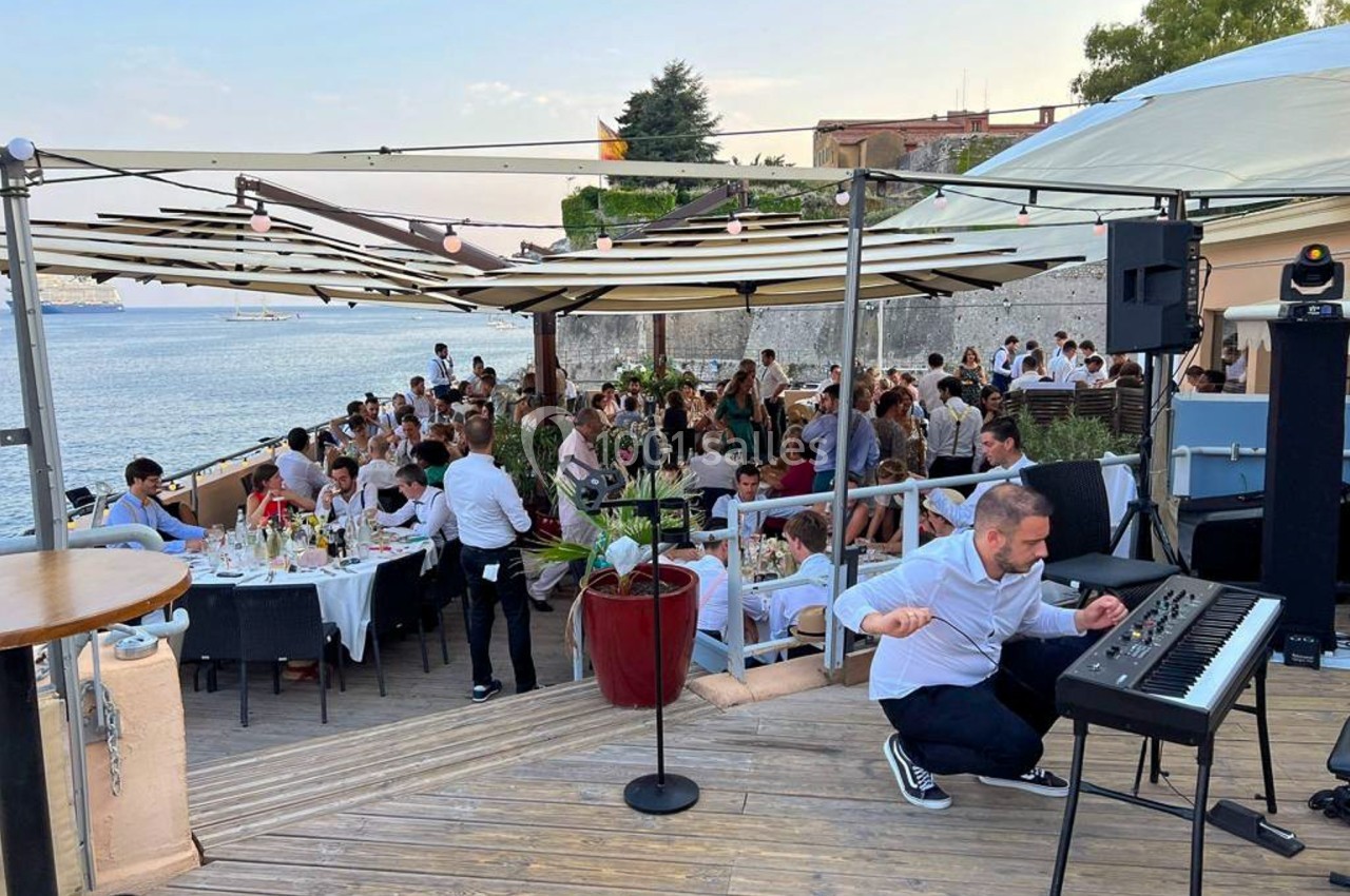Terrasse en bord de mer avec des tables occupées par des convives et un musicien jouant du clavier au premier plan.