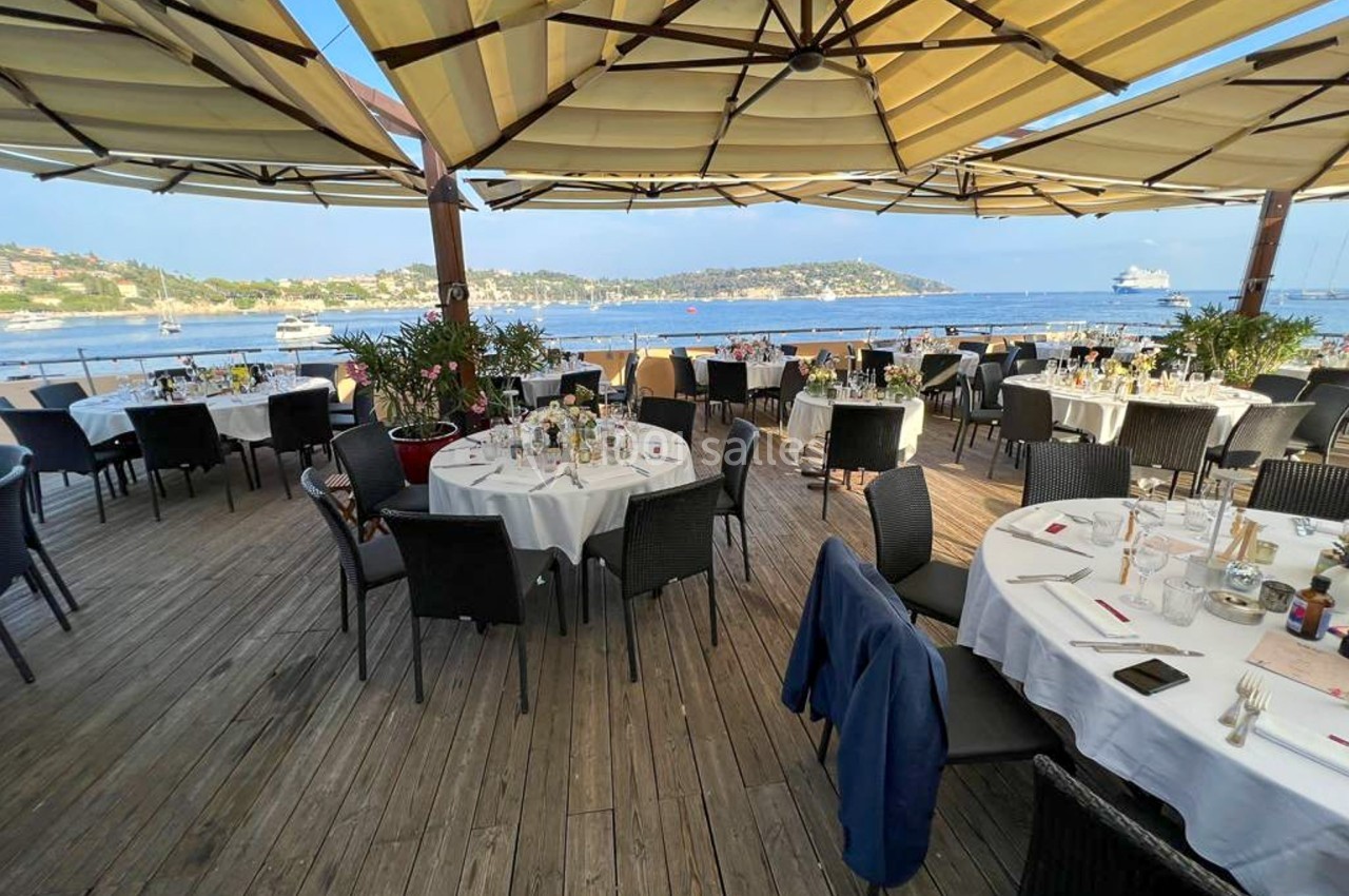 Terrasse d'un restaurant en bord de mer avec tables dressées, vue sur un port et des collines en arrière-plan.