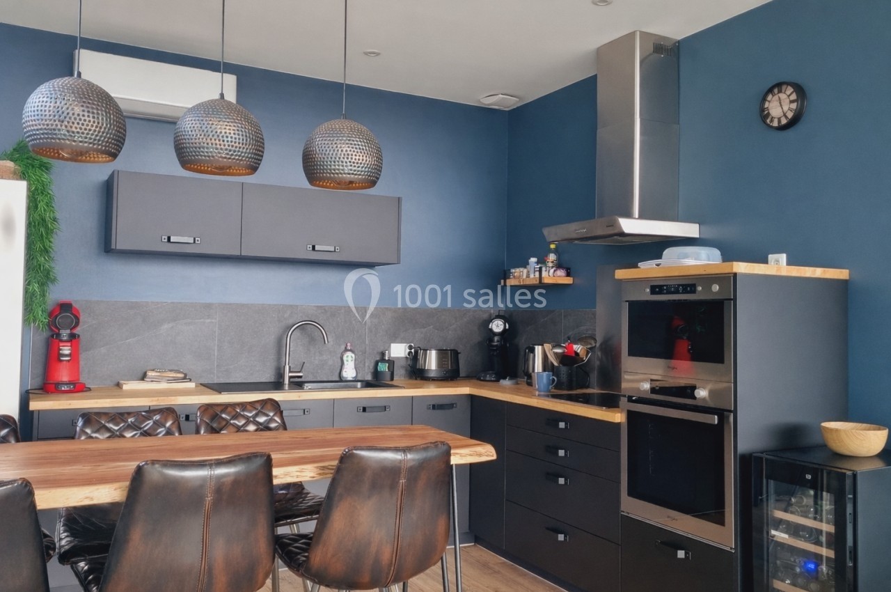 Cuisine moderne avec murs bleus, meubles noirs, plan de travail en bois, électroménagers intégrés et table avec chaises.