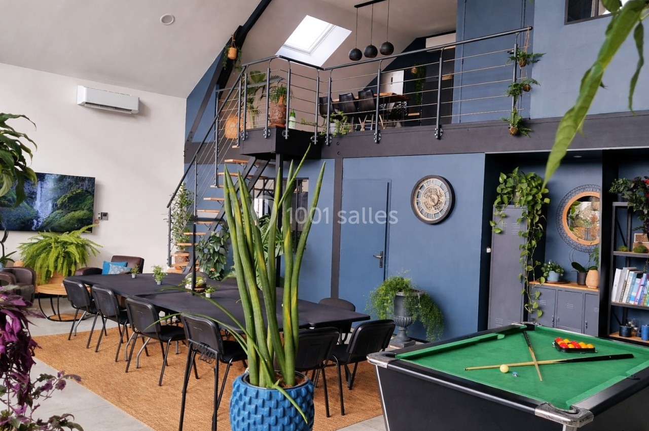 Salle moderne avec mezzanine, table de billard, grande table noire, plantes décoratives et lumière naturelle.