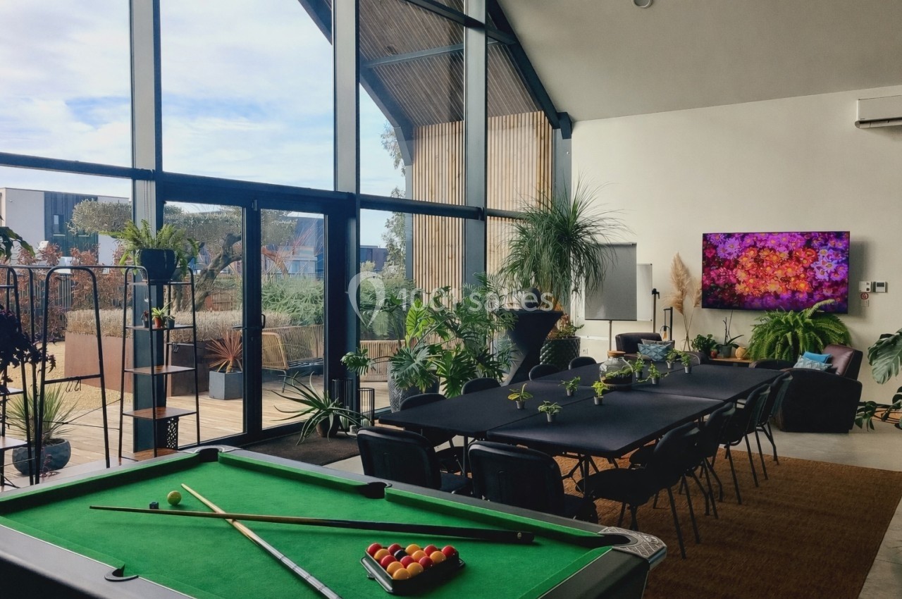 Salle lumineuse avec table de billard, grande table de réunion, plantes et baie vitrée donnant sur une terrasse.