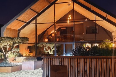 Façade d'une maison moderne en bois et verre, éclairée de nuit, avec jardin et portail en premier plan.