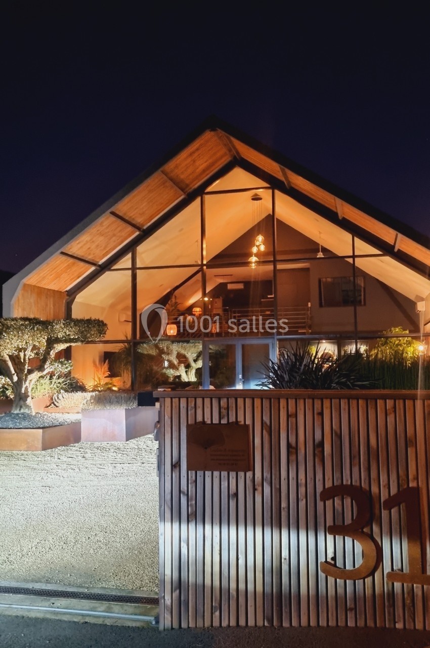 Façade d'une maison moderne en bois et verre, éclairée de nuit, avec jardin et portail en premier plan.