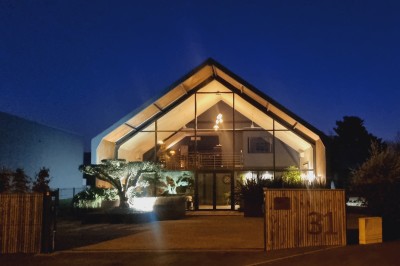 Façade d'une maison moderne en bois et verre, éclairée de nuit, avec jardin et portail en premier plan.