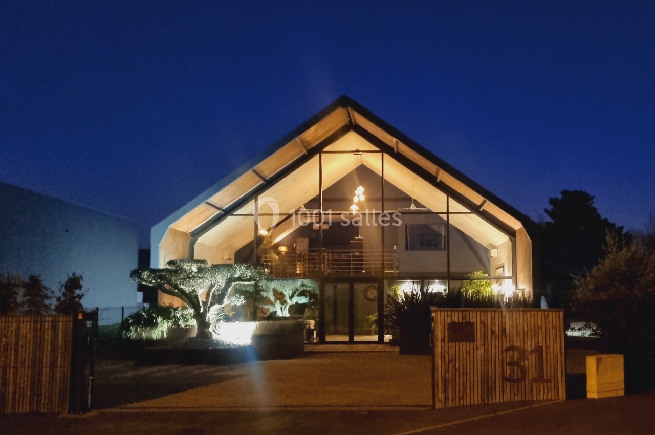 Façade d'un bâtiment moderne avec une grande verrière illuminée, entouré de végétation, photographié de nuit.