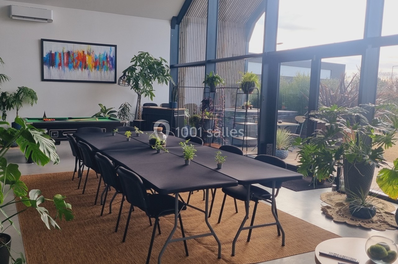 Salle lumineuse avec grande table noire, plantes vertes, billard et baie vitrée donnant sur un extérieur arboré.