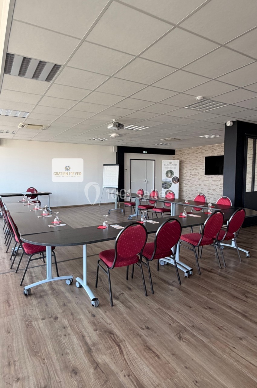 Salle de réunion lumineuse avec tables noires disposées en U, chaises rouges, tableau blanc et projecteur au plafond.