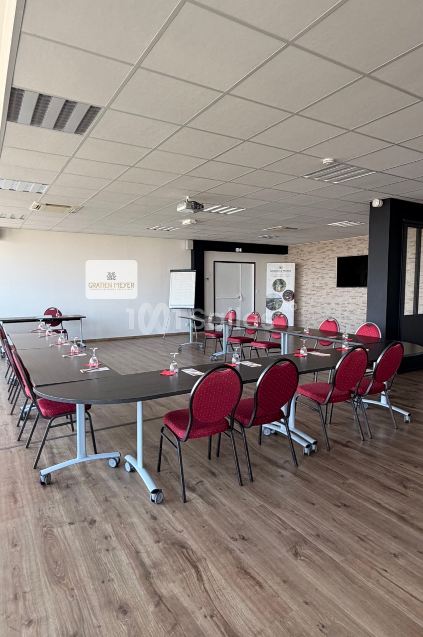 Salle de réunion lumineuse avec tables noires disposées en U, chaises rouges, tableau blanc et projecteur au plafond.