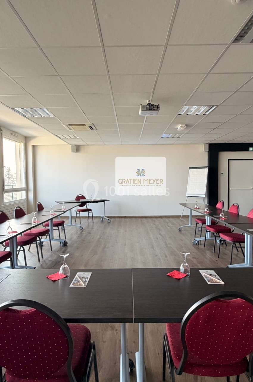 Salle de réunion lumineuse avec tables disposées en U, chaises rouges, écran de projection et paperboard.