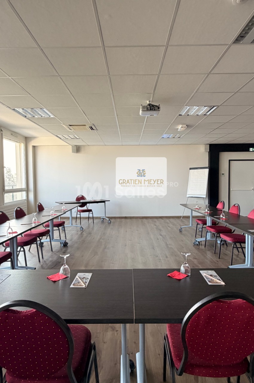 Salle de réunion lumineuse avec tables disposées en U, chaises rouges, écran de projection et paperboard.