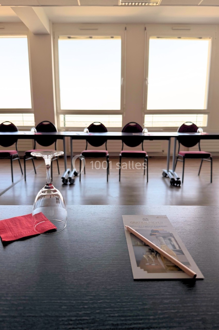 Salle de réunion lumineuse avec des chaises alignées, une table dressée avec un verre, une serviette et un document.